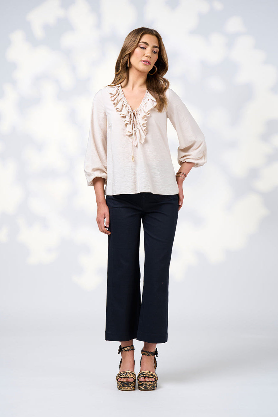 Woman wearing a white blouse with ruffle details and dark pants against a blurred light background