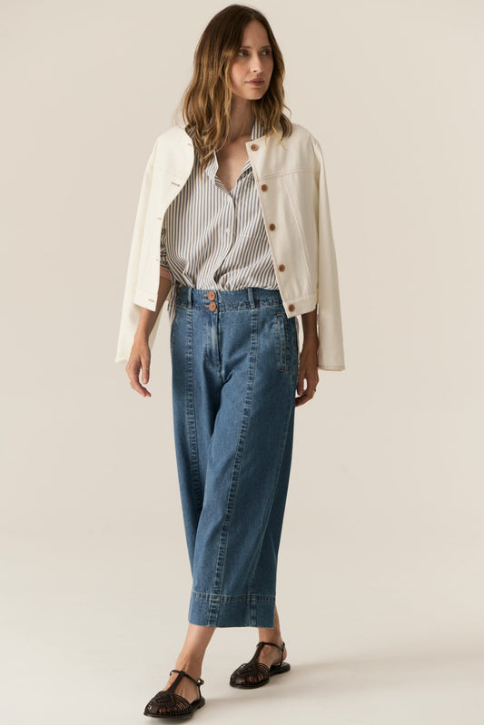 Woman wearing a white jacket, striped shirt, and blue jeans on a beige background