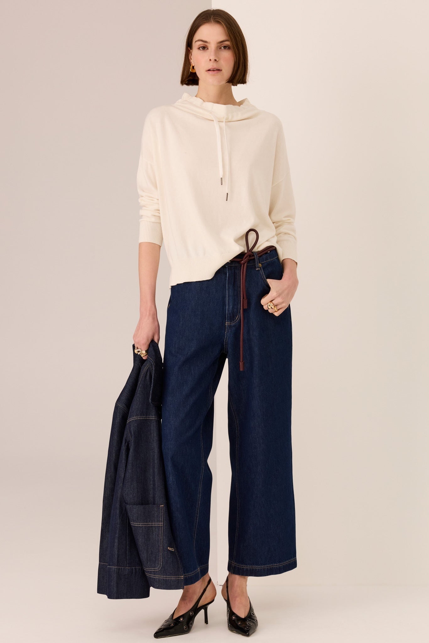 Woman wearing a cream sweater and dark blue wide-leg pants holding a coat on a plain background