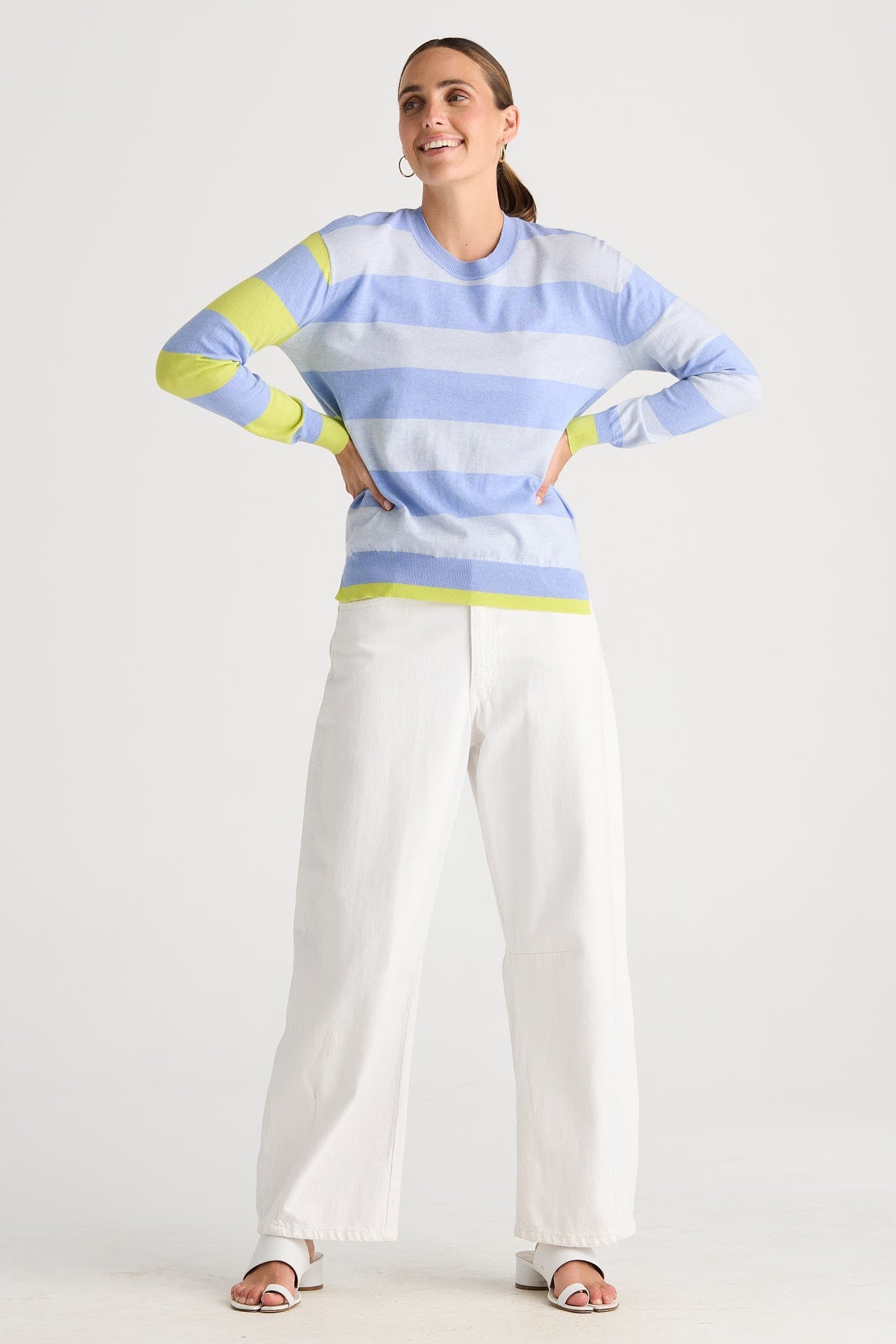 A woman modeling a light blue and white striped long-sleeve knit top with lime green accents on the shoulders, cuffs, and hem, paired with white wide-leg pants and sandals, against a plain light gray studio background.
