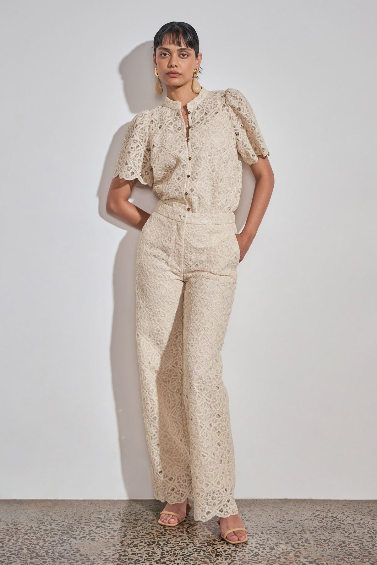 A model stands against a white wall wearing a matching cream lace top and wide-leg pants set, paired with nude flat sandals.