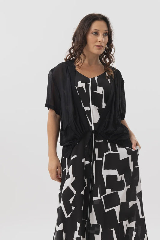 A woman modeling a black sheer draped cardigan over a black and white geometric print dress against a plain light gray studio background.