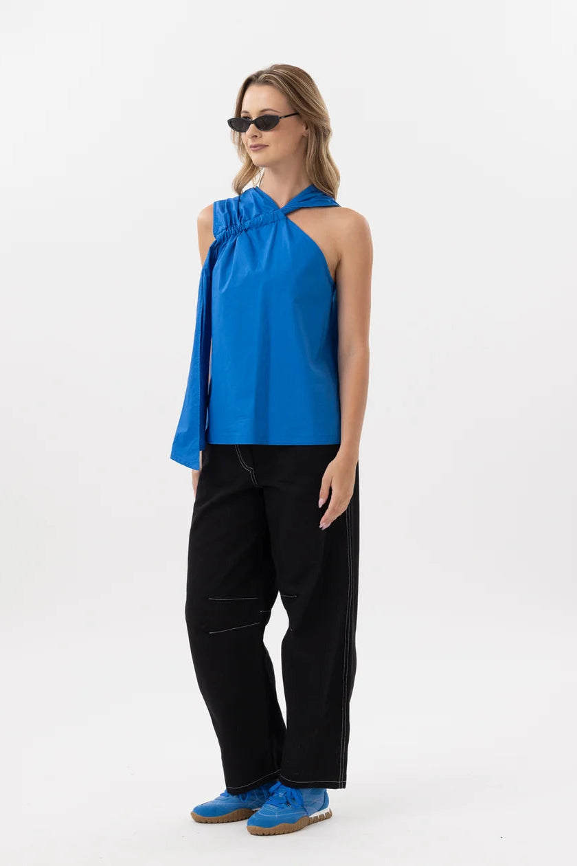 A woman modeling a bright blue halter-style top with a draped sleeve, paired with black pants and blue shoes, photographed against a plain white studio background.