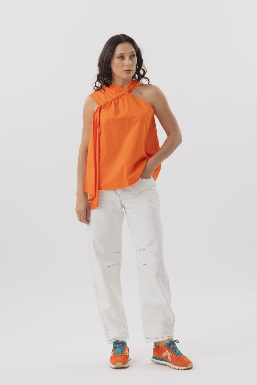 A woman modeling a bright orange halter-neck top with white pants and orange sneakers, standing against a plain light gray studio background.