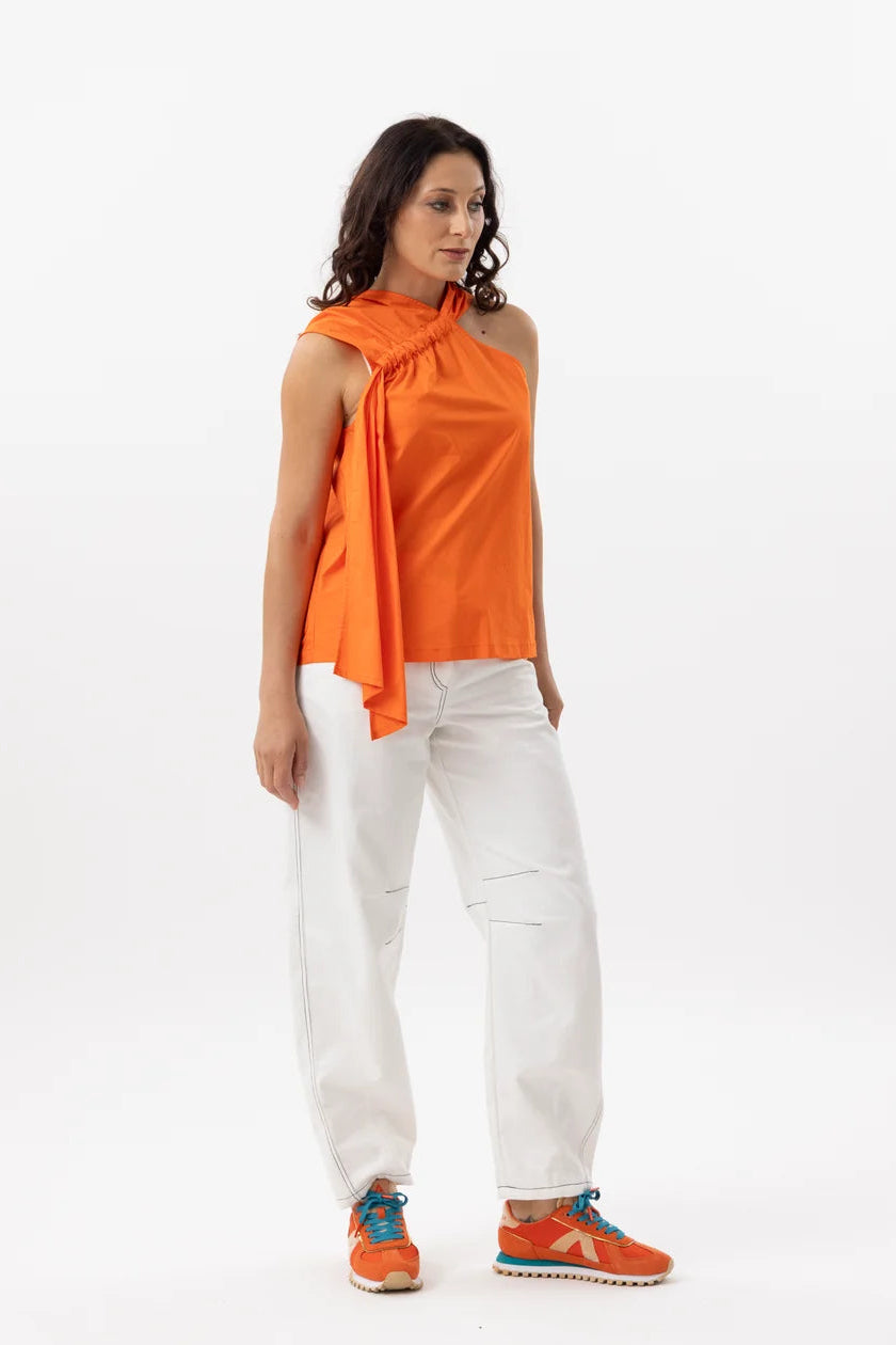 A woman modeling a sleeveless bright orange top with a draped tie detail at the neckline, paired with white pants and orange sneakers, against a plain white studio background.