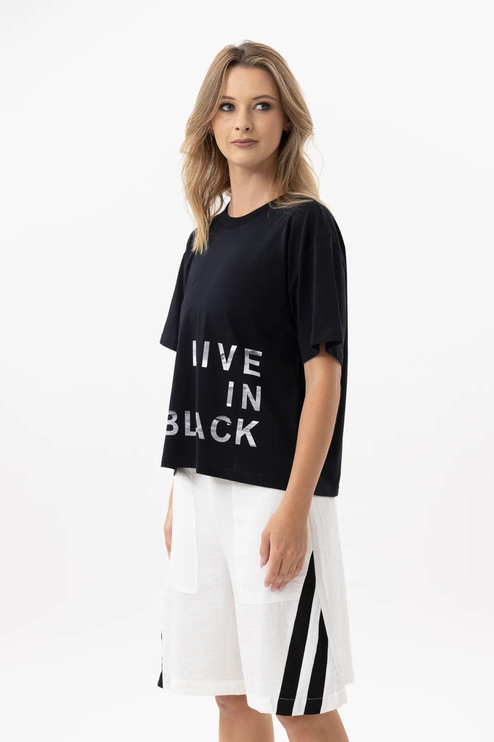 A woman modeling a black oversized t-shirt with white text reading 'LIVE IN BLACK' and white shorts with black side stripes, set against a plain white studio background.