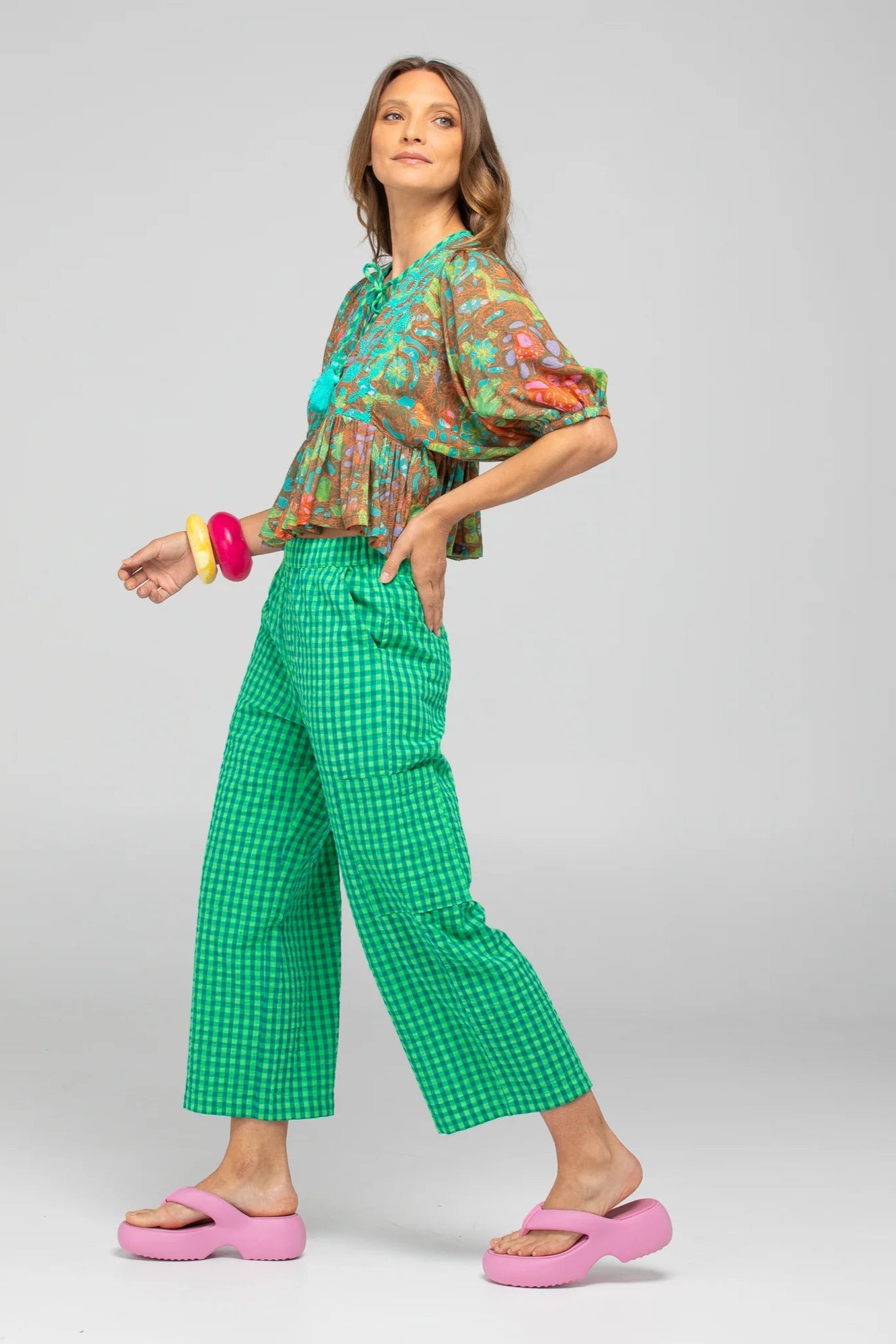 Woman wearing a floral top and green checkered pants with pink shoes on a gray background