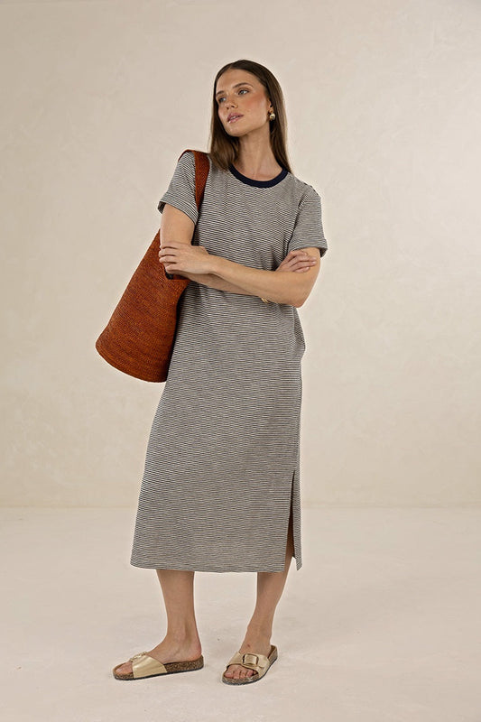 A woman modeling a short-sleeve gray midi dress with a dark navy collar, carrying a large brown woven shoulder bag, and wearing beige slide sandals against a neutral beige studio backdrop.