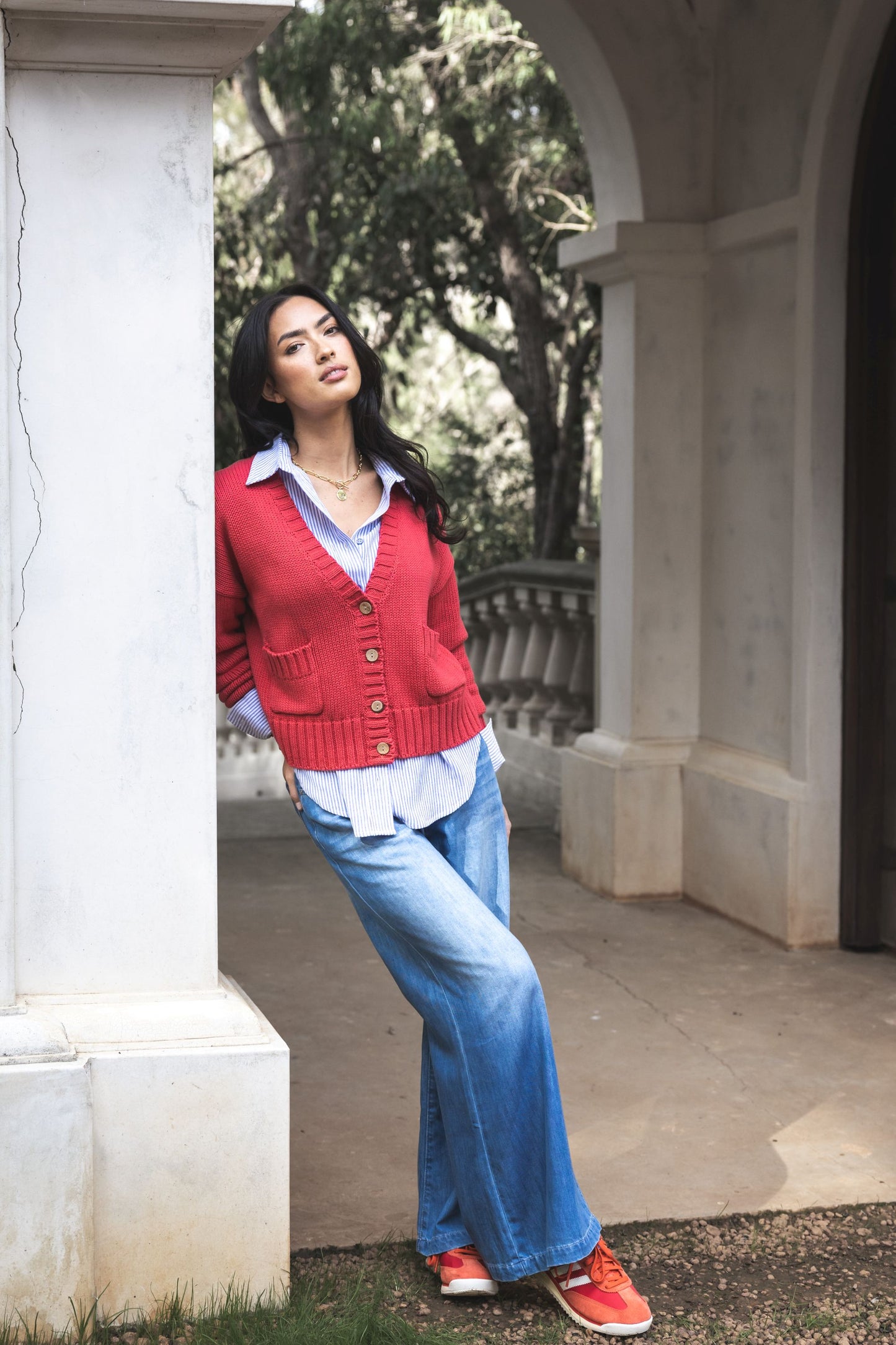 Woman wearing a red cardigan, white shirt, and blue jeans standing in an outdoor setting.