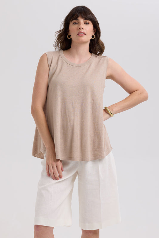 Woman wearing a beige sleeveless top and white pants on a plain background