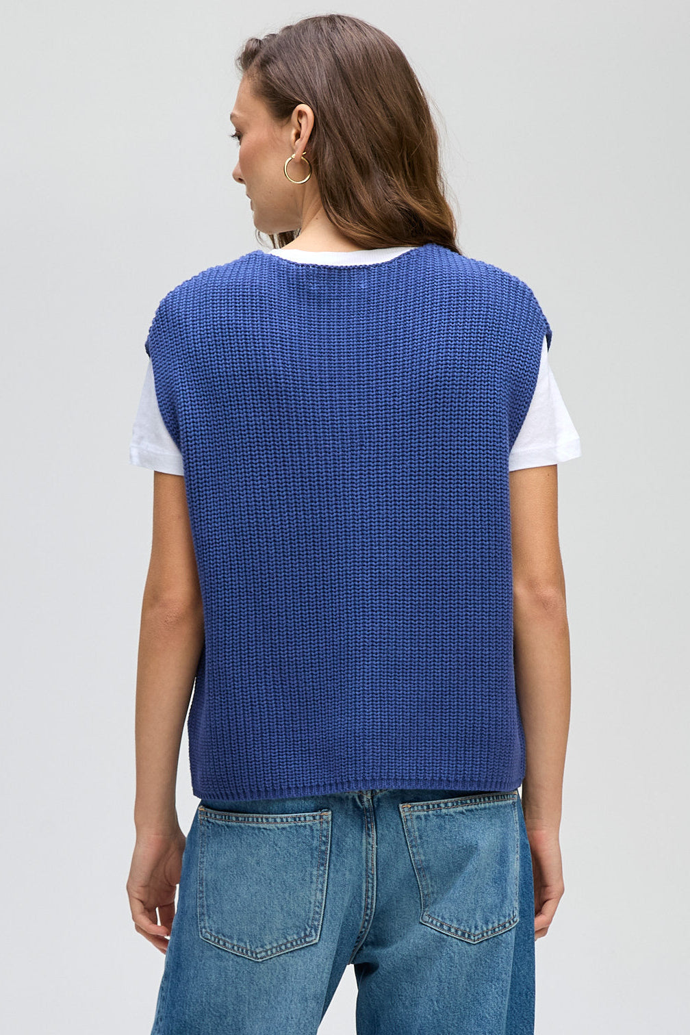 Person wearing a blue knitted vest over a white shirt with blue jeans on a light gray background