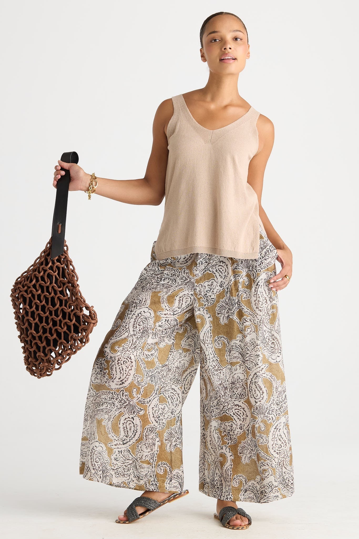 A woman modeling a beige sleeveless tank top and wide-leg pants with a brown, white, and gray paisley pattern. She is holding a brown woven mesh bag against a plain light gray studio background.