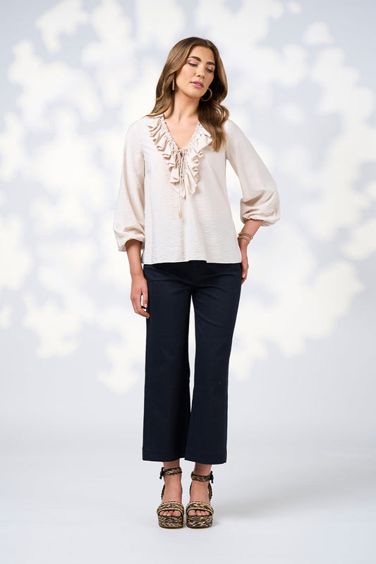 Woman wearing a white blouse with ruffle details and dark pants against a blurred light background