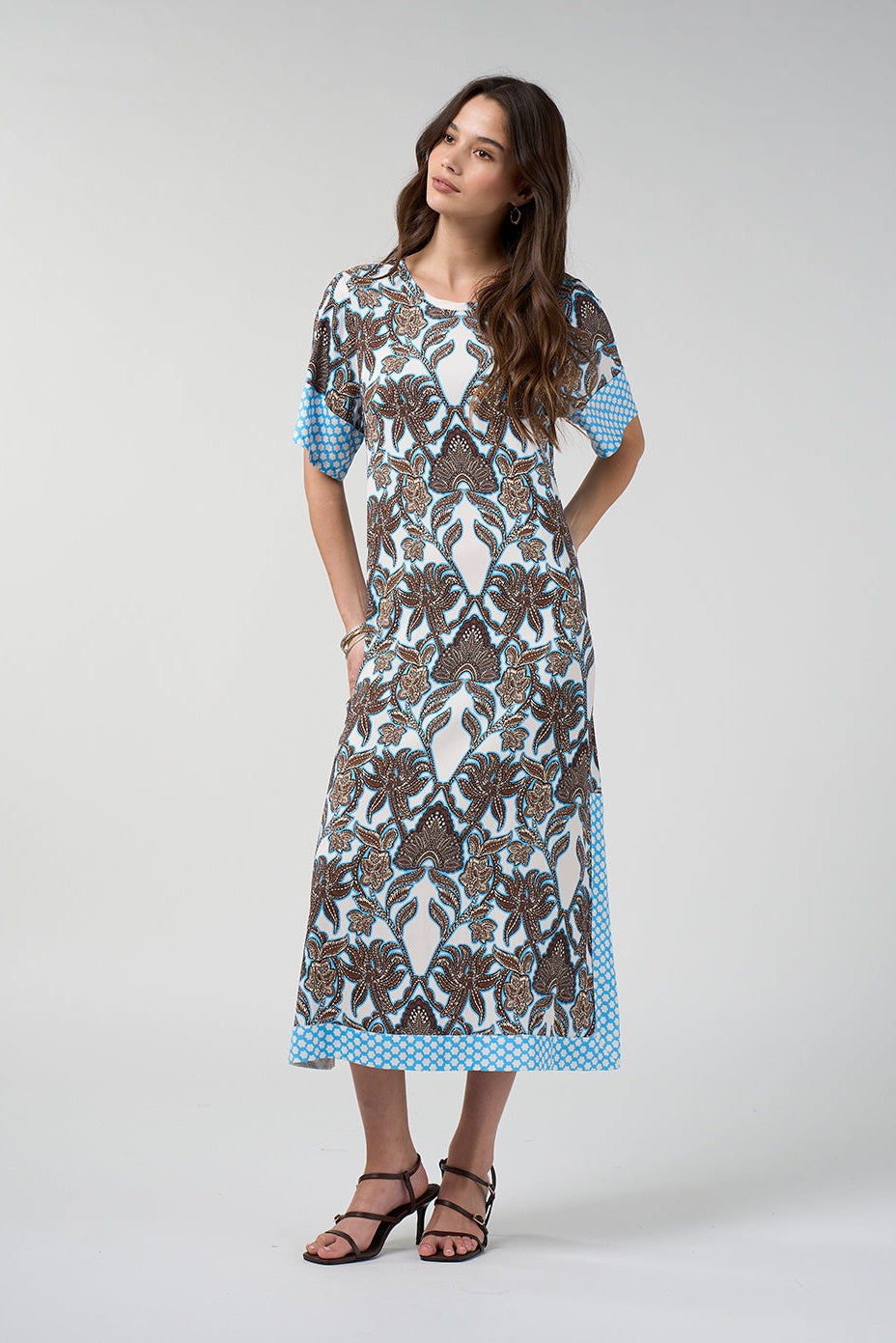A woman modeling a short-sleeved midi dress with a brown and blue floral batik pattern on a white background, featuring light blue and white polka dot trim on the sleeves and hem, paired with black strappy sandals against a light gray studio background.