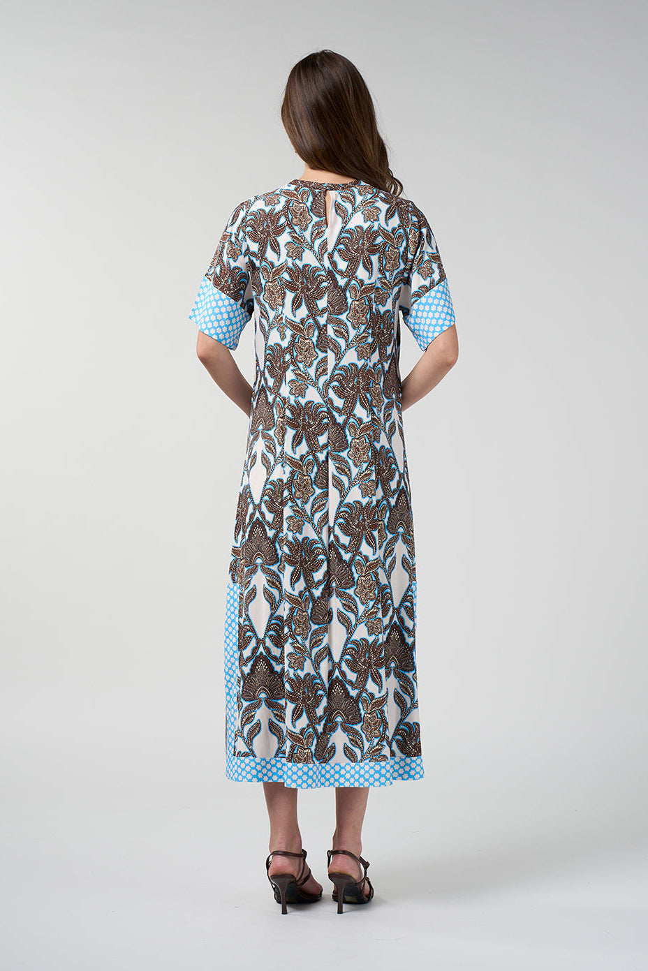 A woman viewed from the back wearing a knee-length dress with a brown and blue floral print on a white base, featuring light blue polka-dot trim at the sleeve cuffs and hem, paired with black heeled sandals against a light gray studio background.