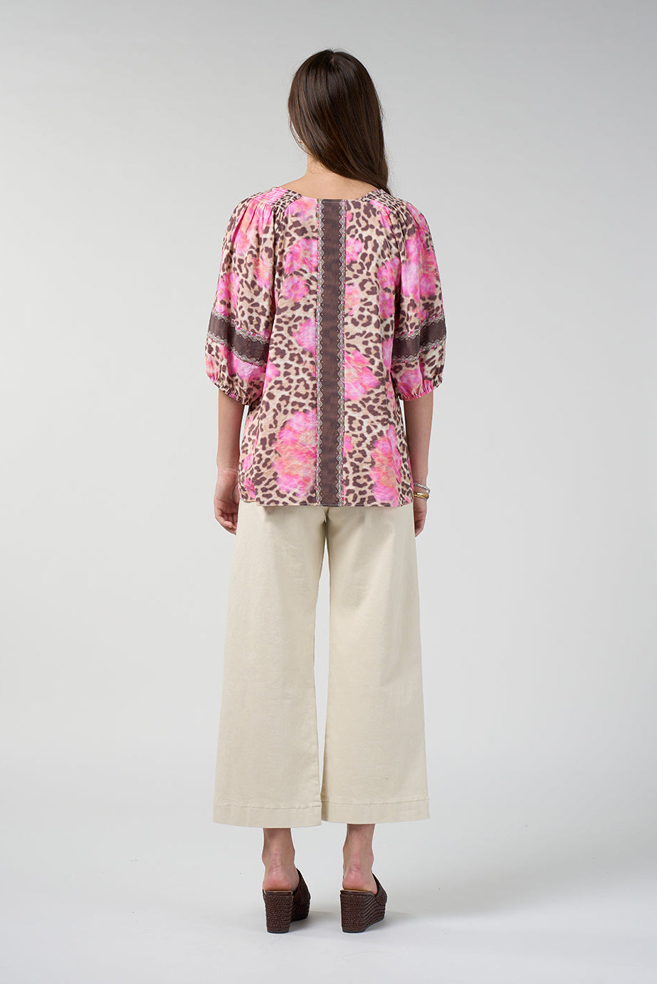 A woman viewed from the back wearing a pink and leopard-print blouse with three-quarter sleeves and cream wide-leg cropped pants, standing against a light gray studio background.