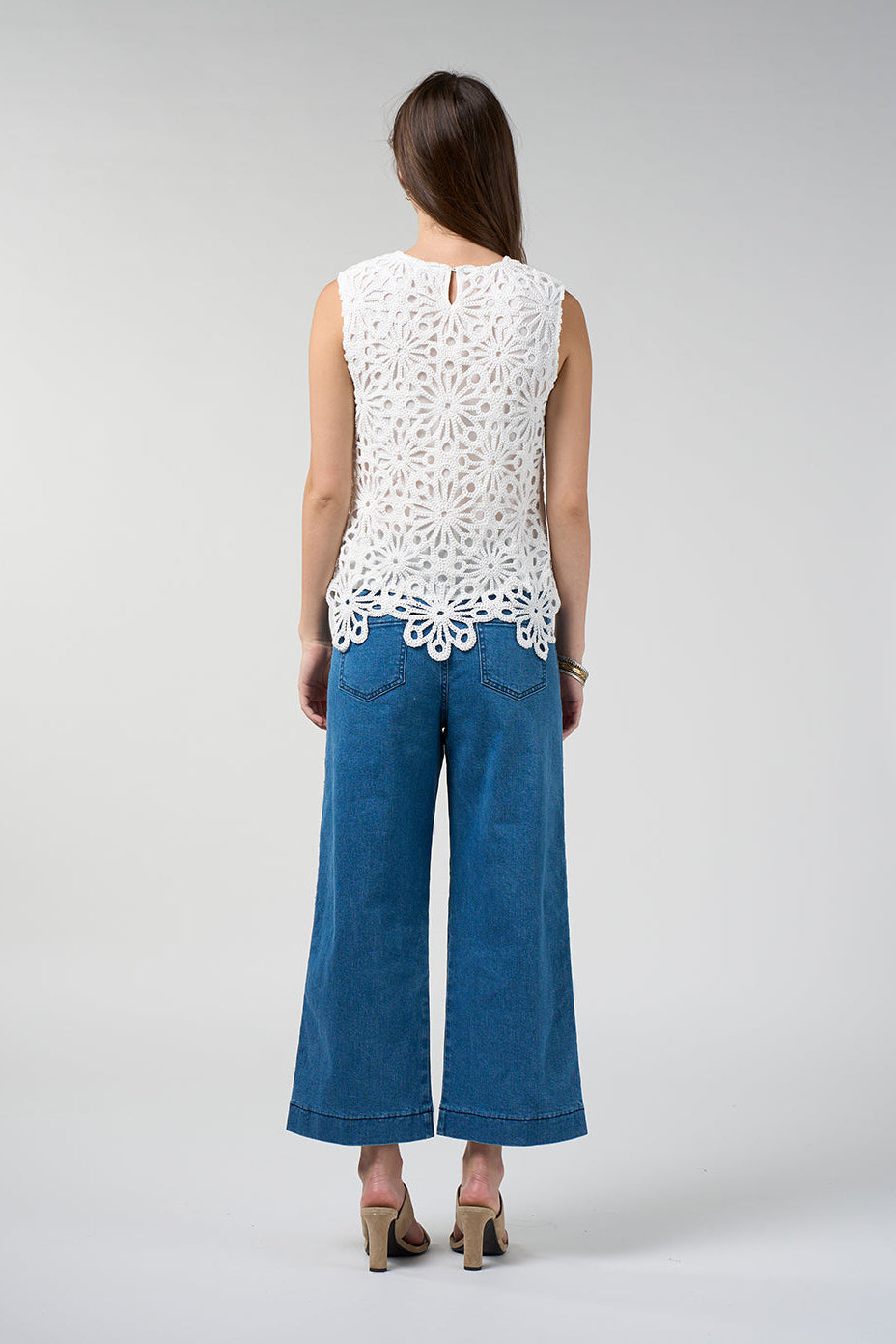 A woman viewed from the back wearing a white sleeveless crochet lace top and blue wide-leg jeans, standing against a light gray studio background.