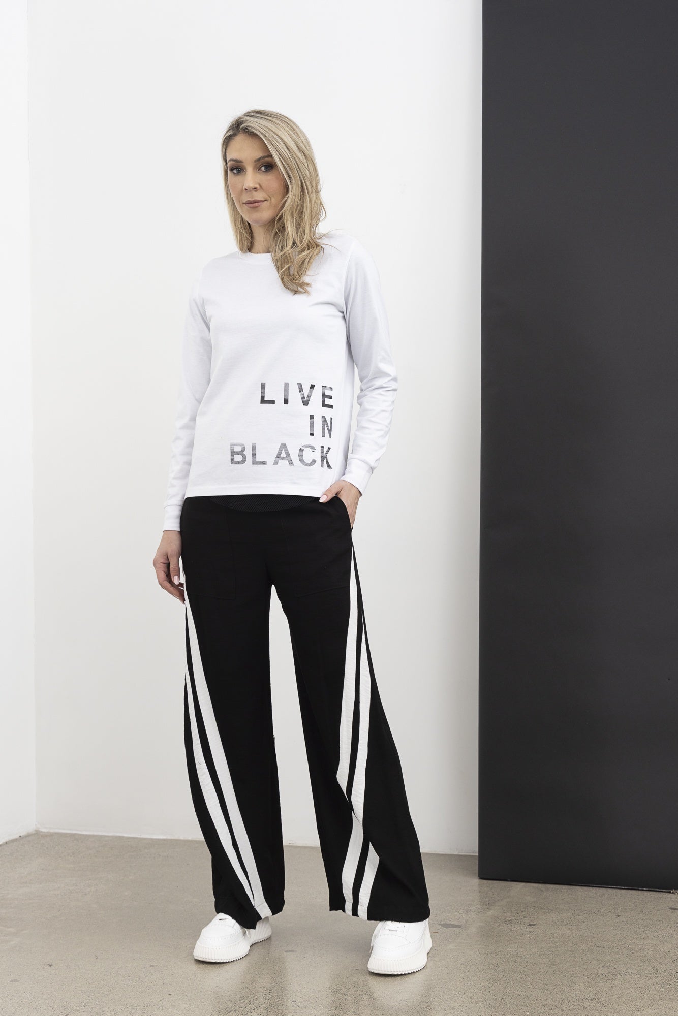Woman wearing a white sweatshirt with text and black pants with white stripes, standing against a white wall.