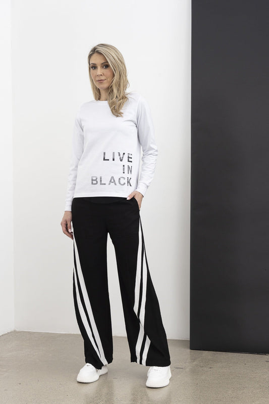 Woman wearing a white sweatshirt with text and black pants with white stripes, standing against a white wall.
