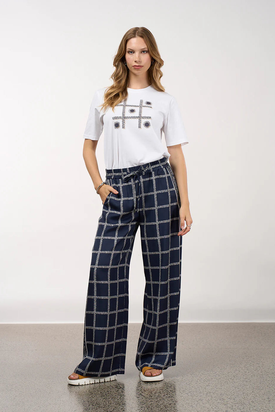 Woman wearing a white t-shirt with a graphic design and navy checkered pants on a plain background