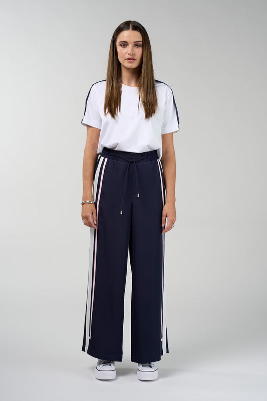 A woman stands against a light gray studio background wearing a white short-sleeve T-shirt with dark blue sleeve stripes, navy wide-leg drawstring pants with white side stripes, and white sneakers.