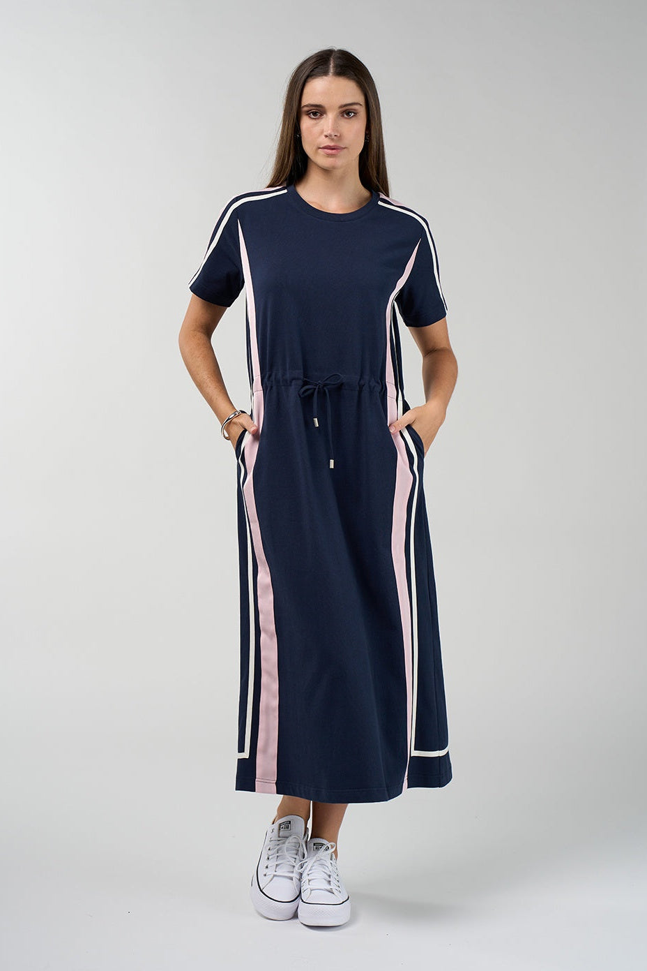 A woman modeling a navy blue short-sleeve maxi dress with pink and white side stripes, a drawstring waist, and side pockets, paired with white sneakers against a light gray studio background.