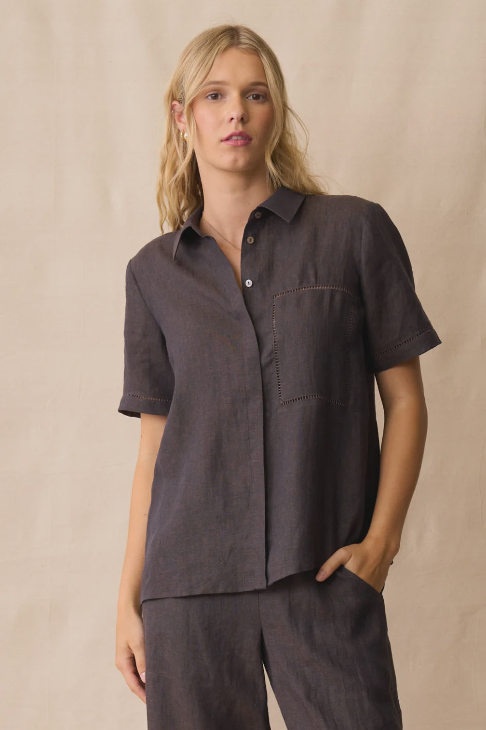 A woman modeling a dark brown short-sleeve button-up shirt and matching pants against a neutral beige backdrop.