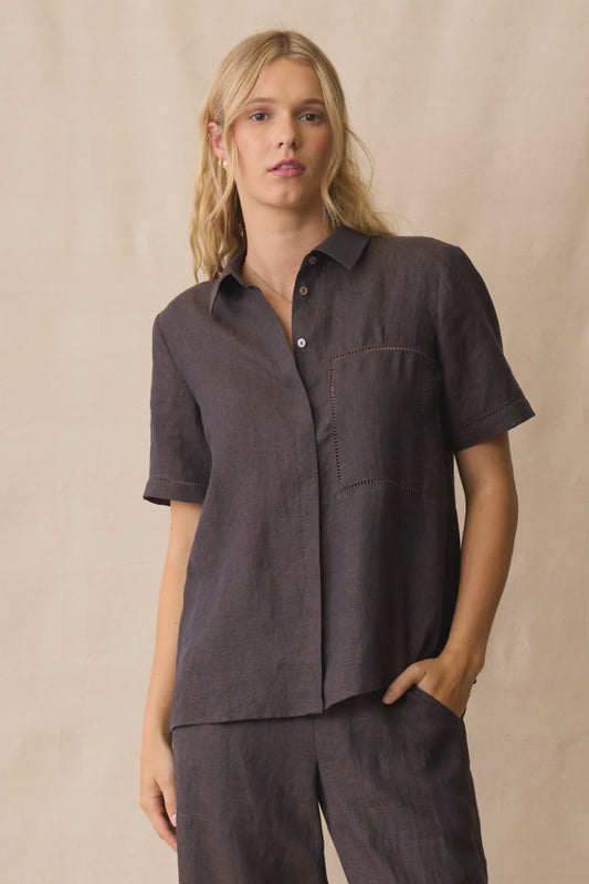A woman modeling a dark brown short-sleeve button-up shirt and matching pants against a neutral beige backdrop.