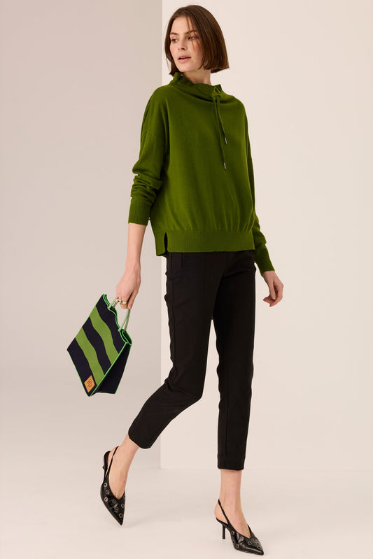 Woman wearing a green sweater and black pants holding a green and black striped bag on a beige background