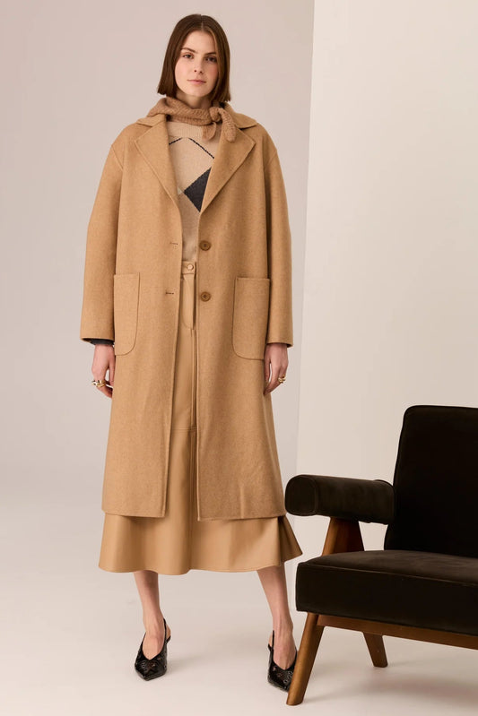 Woman wearing a long beige coat standing next to a black chair on a plain background