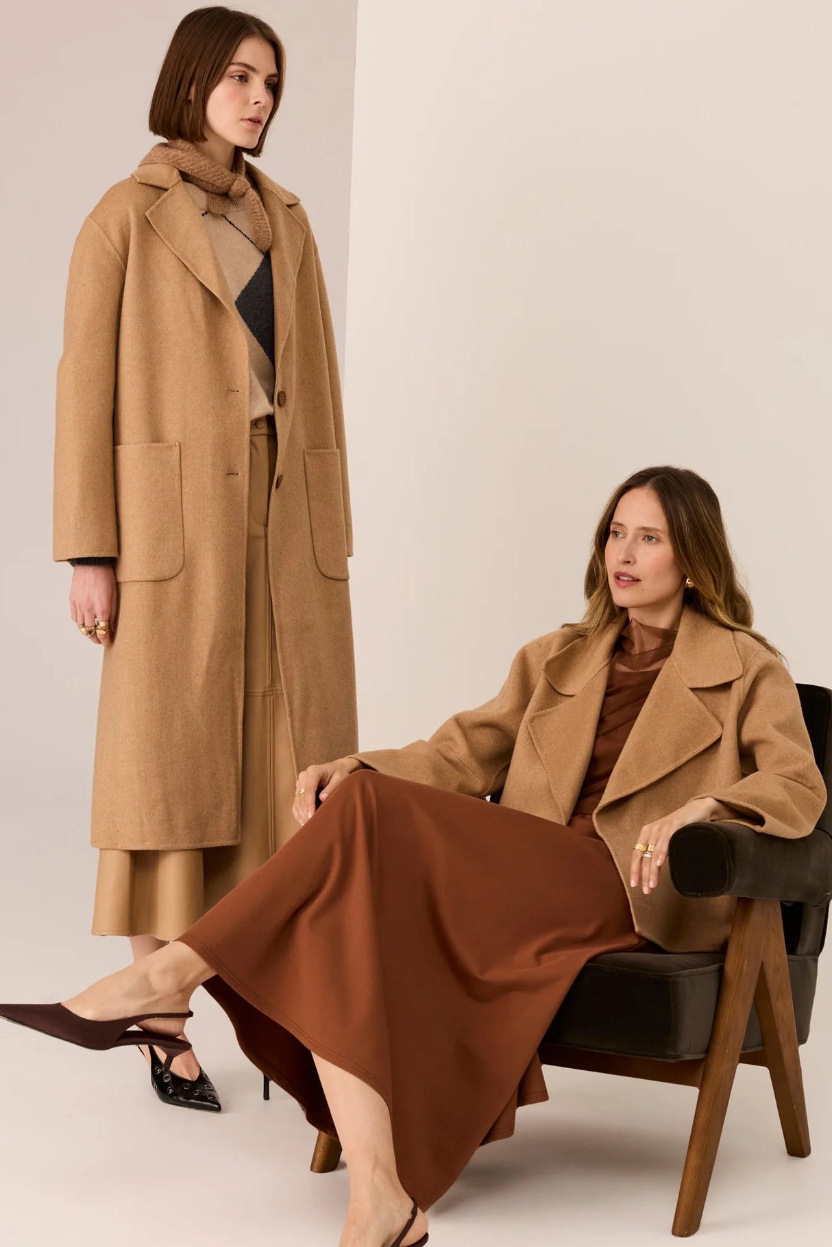 Two women wearing brown coats against a plain background