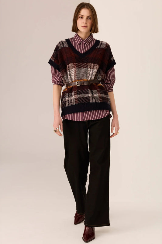 Woman wearing a patterned sweater vest over a striped shirt with black pants on a beige background