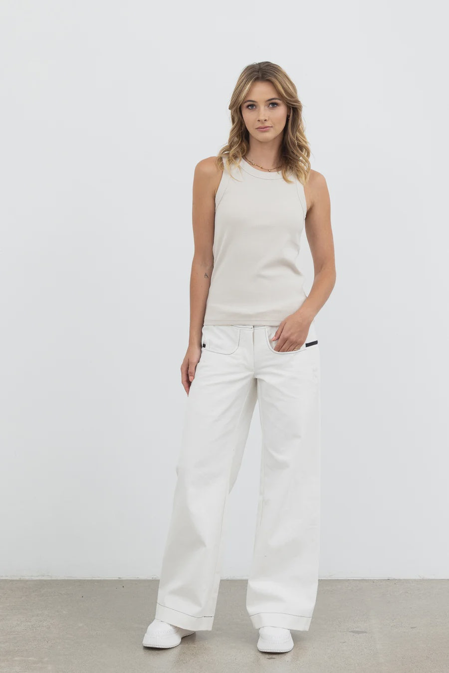 A woman modeling a sleeveless cream knit top and matching white wide-leg pants against a plain light gray studio wall.