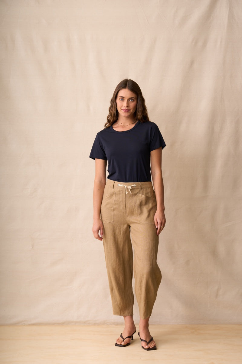 A woman stands against a beige fabric backdrop wearing a dark navy short-sleeve t-shirt, tan cropped drawstring pants, and black sandals.