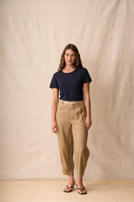 A woman stands against a beige fabric backdrop wearing a dark navy short-sleeve t-shirt, tan cropped drawstring pants, and black sandals.