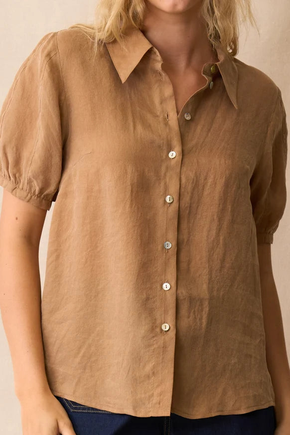 A woman wearing a tan short-sleeve linen button-up shirt with puff sleeves and white buttons, paired with dark blue jeans, against a neutral beige background.