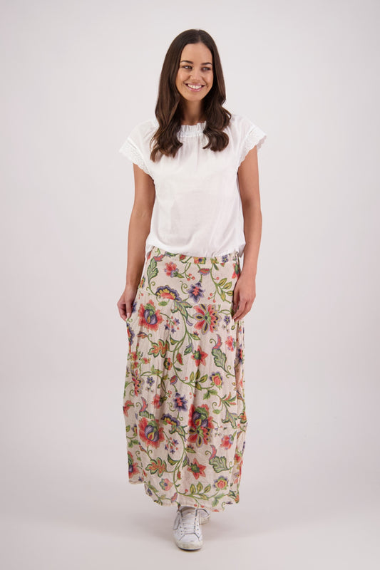 A woman modeling a white short-sleeve blouse and a long cream maxi skirt with a colorful floral print, standing against a light gray studio background.