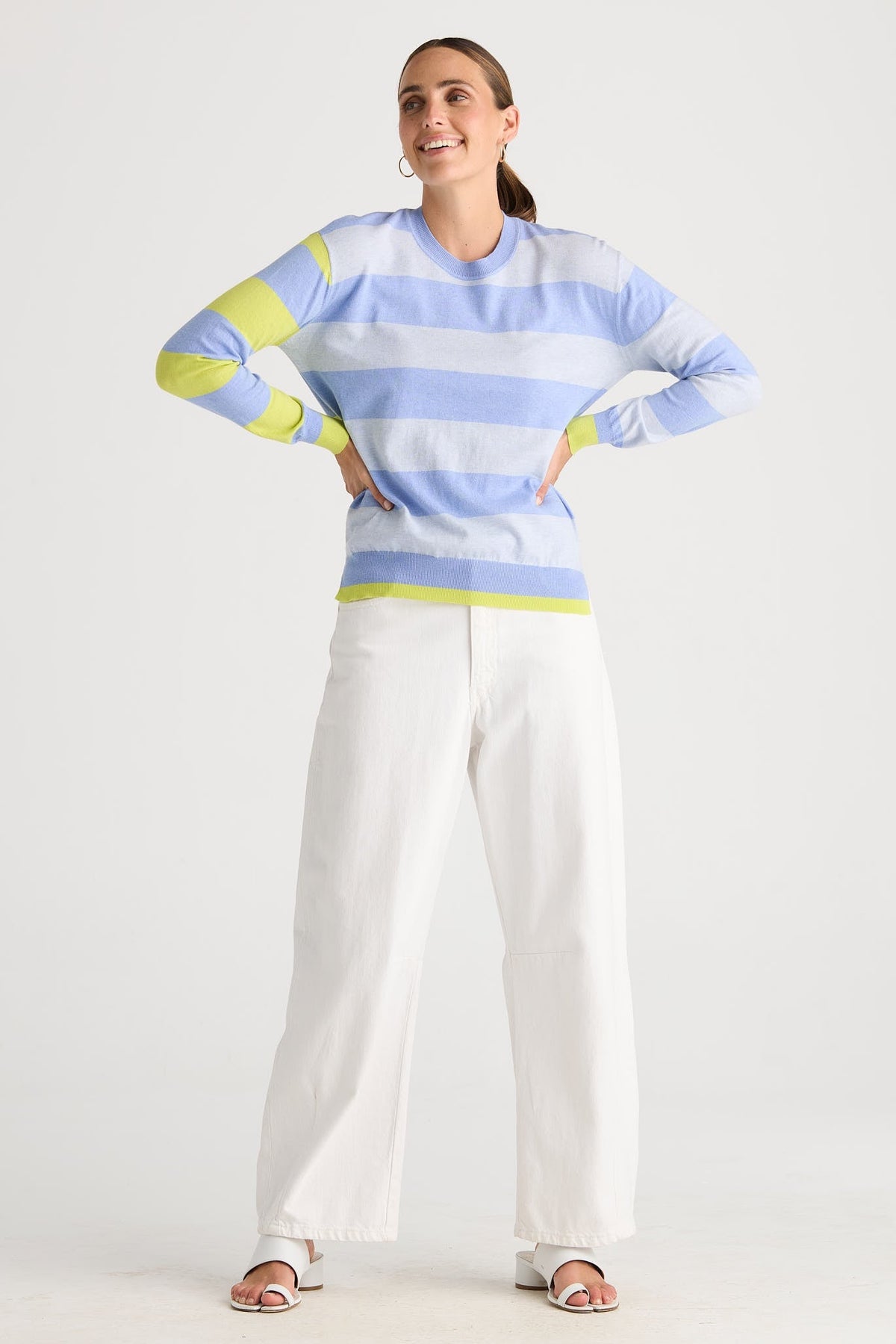 A woman modeling a light blue and white striped long-sleeve knit top with lime green accents on the shoulders, cuffs, and hem, paired with white wide-leg pants and sandals, against a plain light gray studio background.