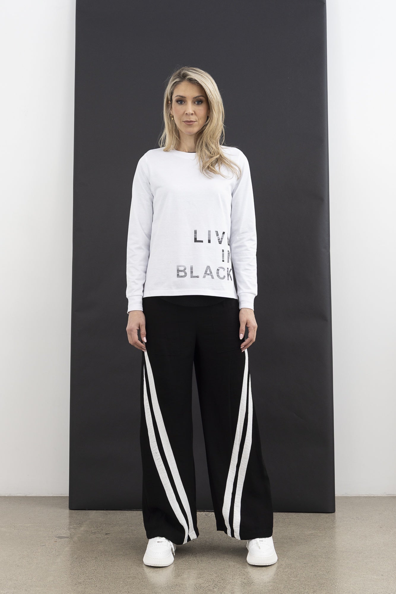 Woman wearing a light gray sweatshirt with text and black pants with white stripes, standing against a black and white background.