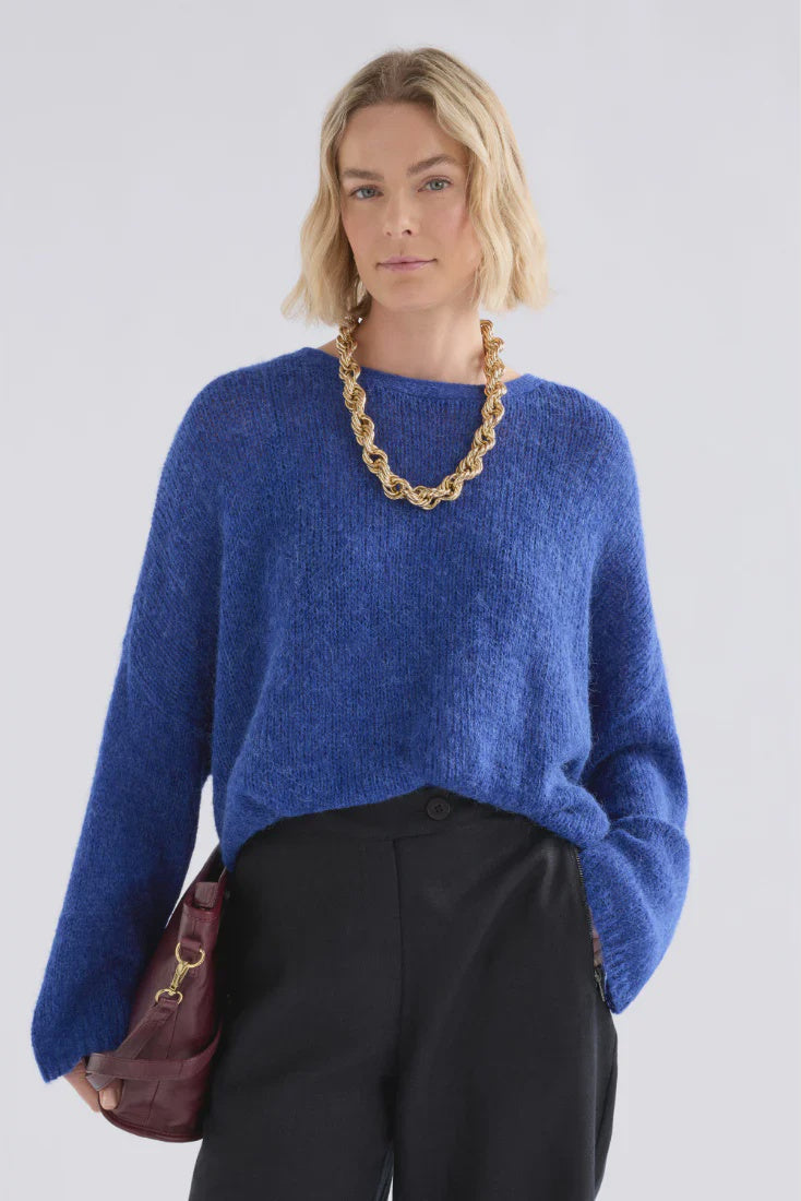 Woman wearing a blue sweater and black pants with a gold chain necklace against a white background